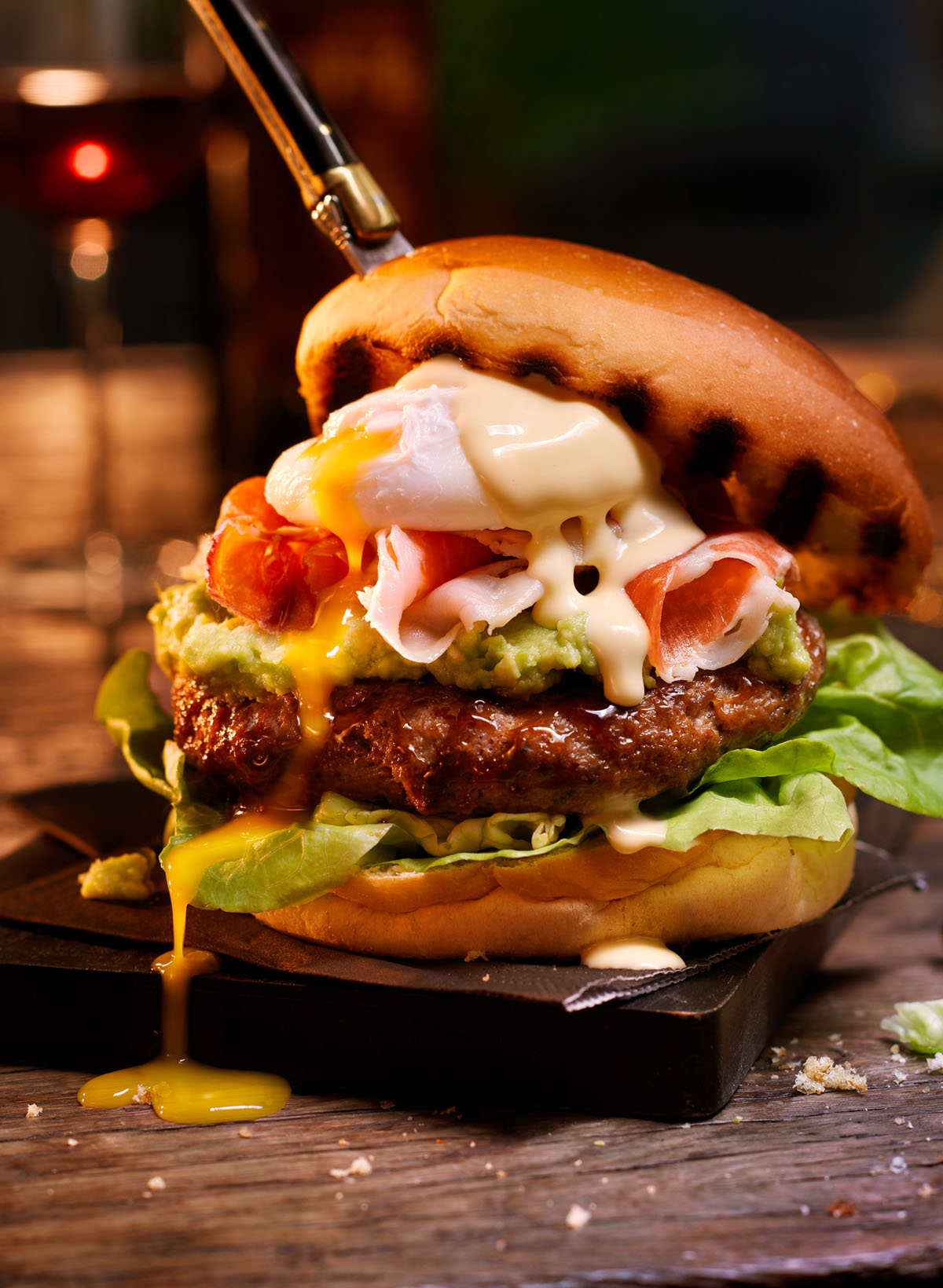 Picture of delicious hamburger with lettuce, beef patty, guacamole, prosciutto, poached egg with yolk oozing out and sauce on top with a knife sticking through the top bun in a ambiance setting of dark wood and black cutting board with blurry background