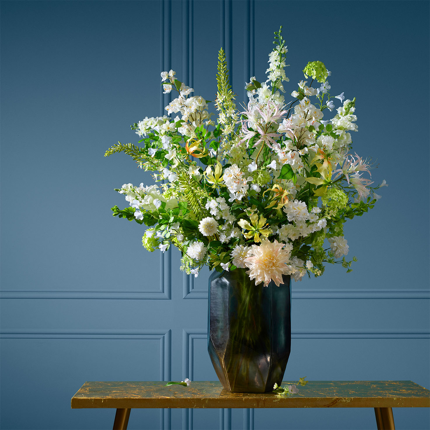 Groot boeket van witte zijden bloemen in een donkere vaas, gefotografeerd tegen een elegante blauwe muur. Het arrangement bevat diverse bloemensoorten met groene accenten, wat het geheel een frisse en verfijnde uitstraling geeft.