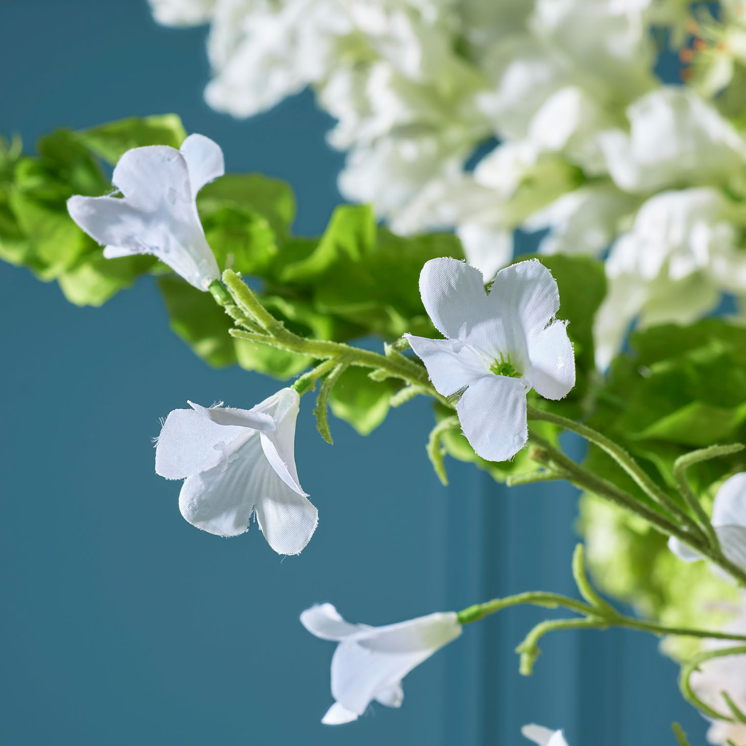 Close-up van delicate witte zijden bloemen met groene bladeren, gefotografeerd tegen een zachte blauwe achtergrond. De bloemen zijn gedetailleerd weergegeven, waardoor hun textuur en natuurlijke uitstraling goed tot hun recht komen.