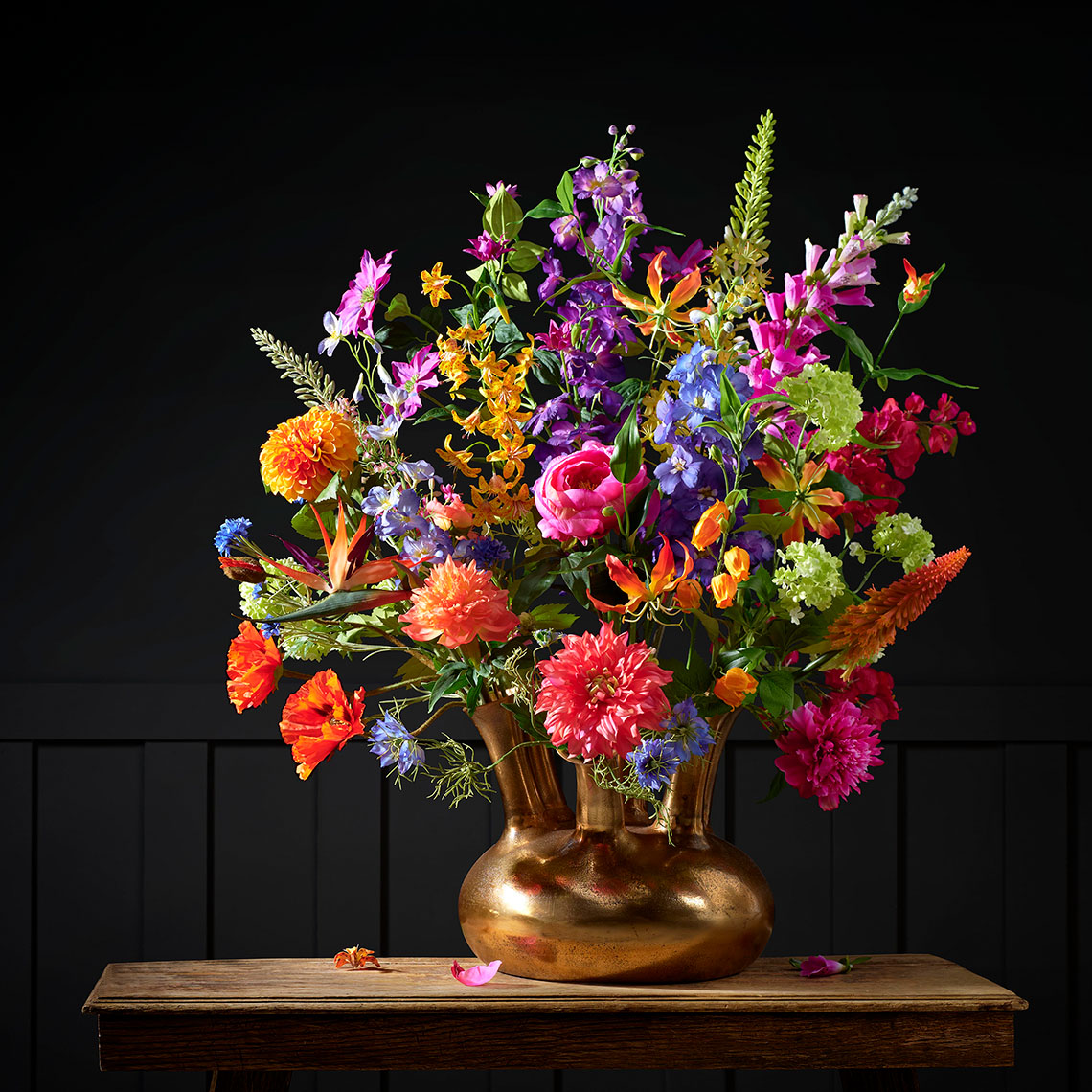 Groot kleurrijk boeket van zijdebloemen in een gouden tuit vaas, gefotografeerd tegen een elegante donker grijze muur. Het arrangement bevat diverse bloemensoorten met allerlei kleuren, wat het geheel de uitstraling geeft van een explosie van keuren.