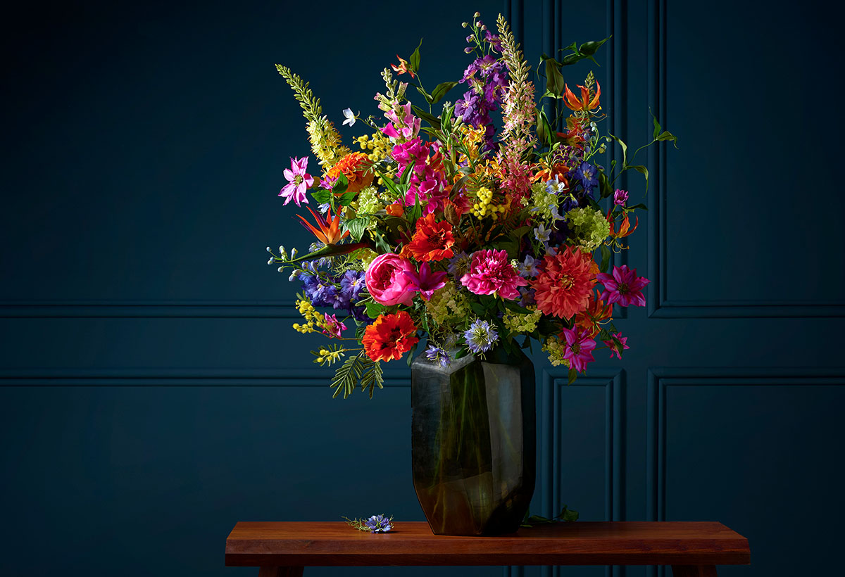 A colorful, classic floral arrangement of silk flowers in a vase, influenced by the still lifes of the Dutch Masters. The arrangement is professionally photographed against a dark background, enhancing the vibrant colors and details of the flowers.