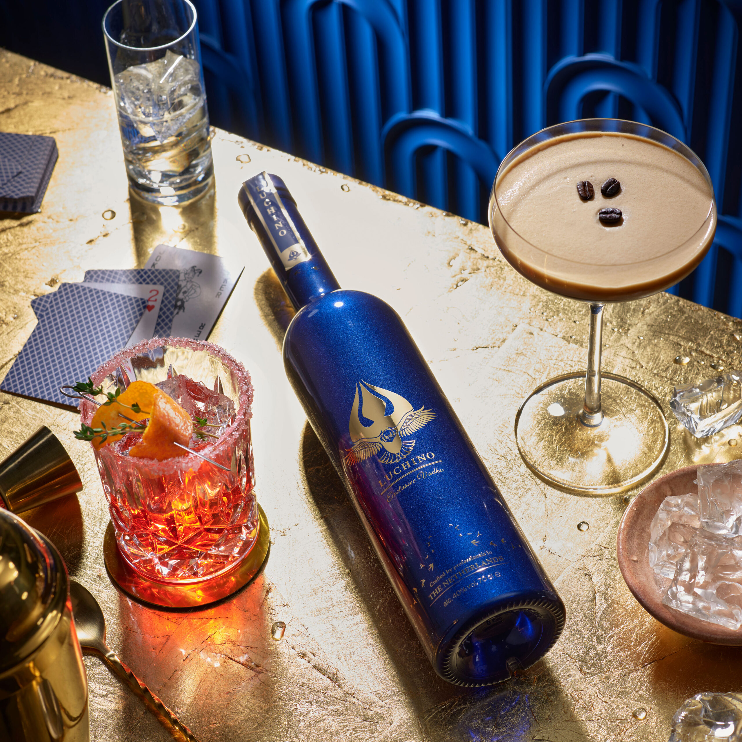 A vodka bottle surrounded by cocktails in a luxurious setting on a golden table against a blue backdrop.